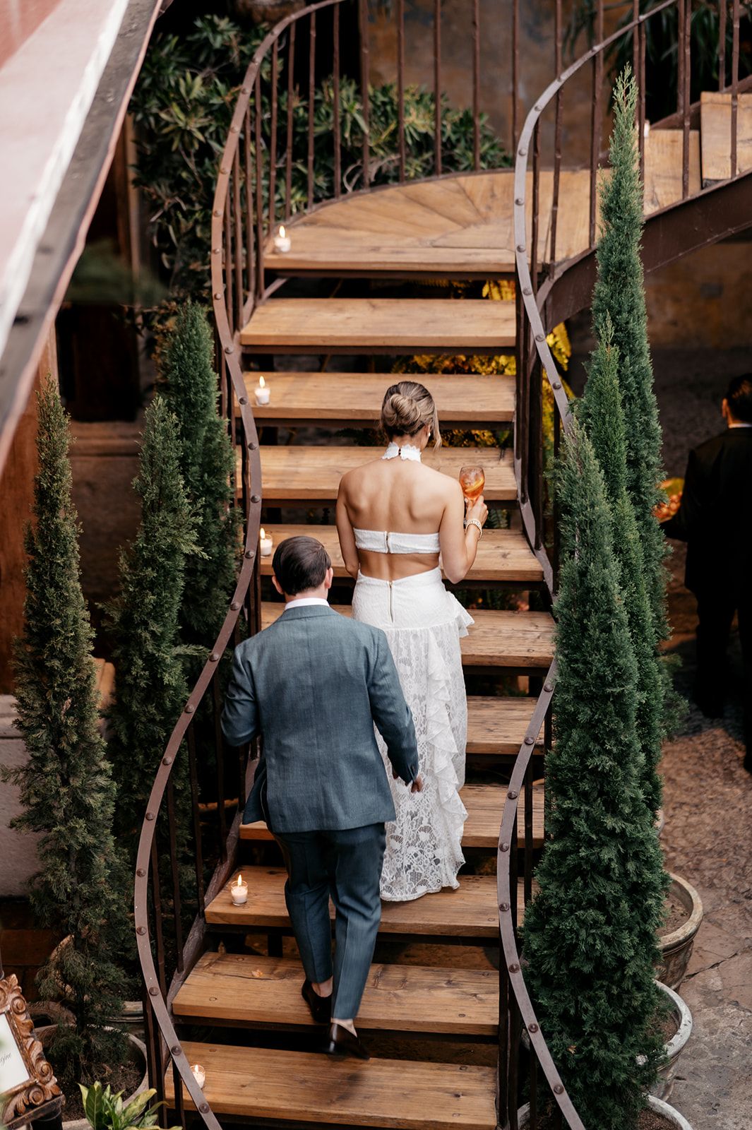 Bride and groom ascending a curved wooden staircase surrounded by tall cypress trees at an elegant wedding venue.