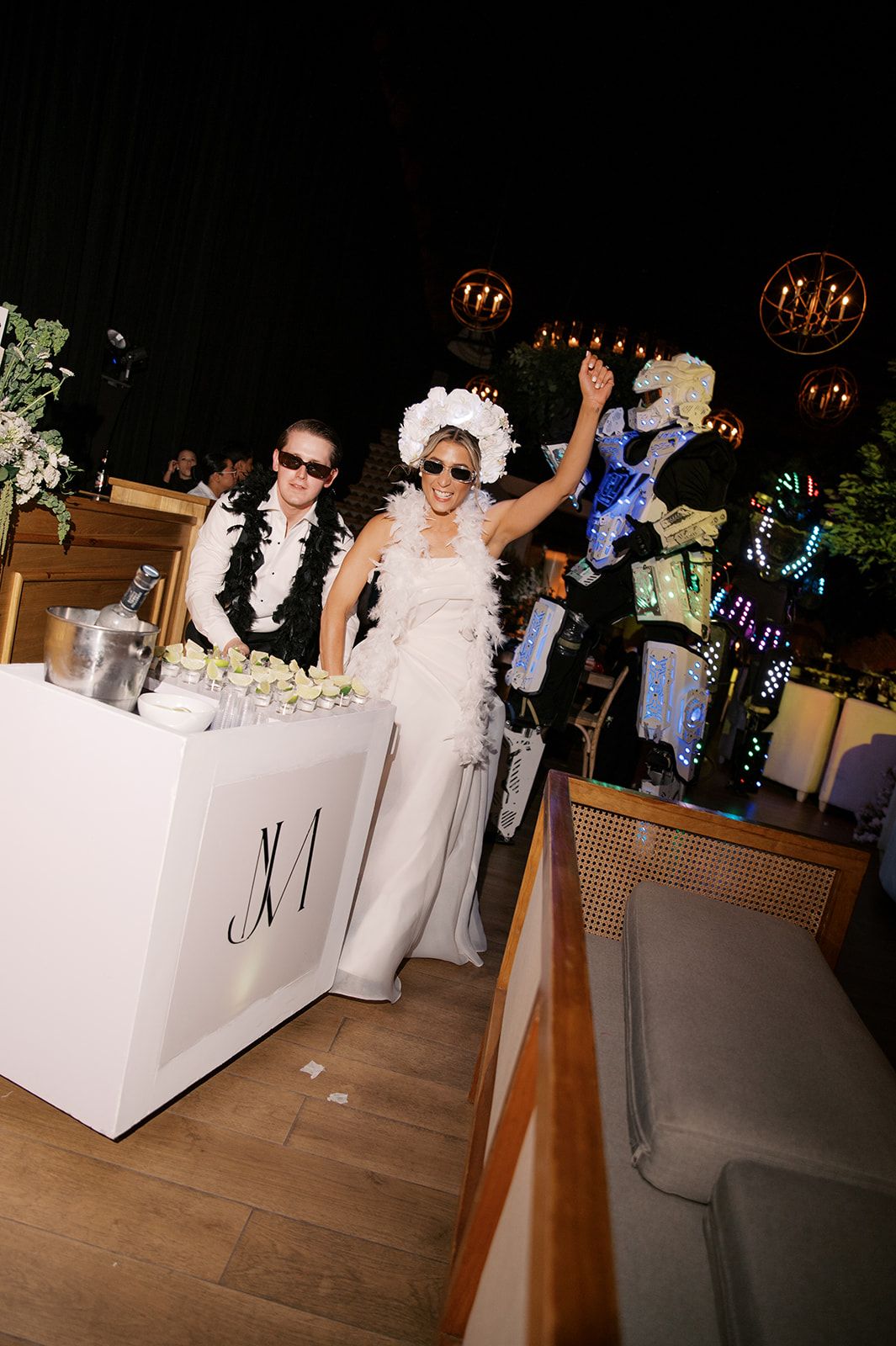 Bride and groom in sunglasses celebrate at wedding reception DJ booth with white floral decor and festive lighting.