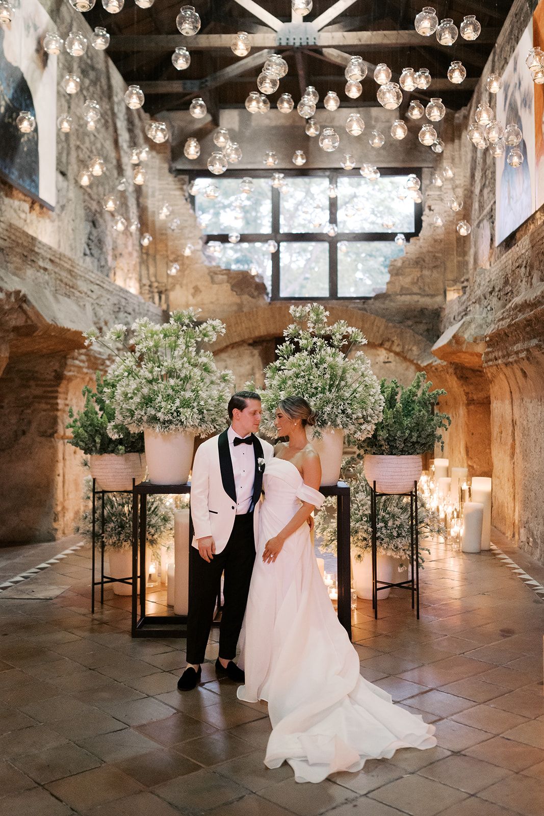 Elegant wedding couple embracing under globe lights in a rustic stone venue with white floral arrangements and candles.