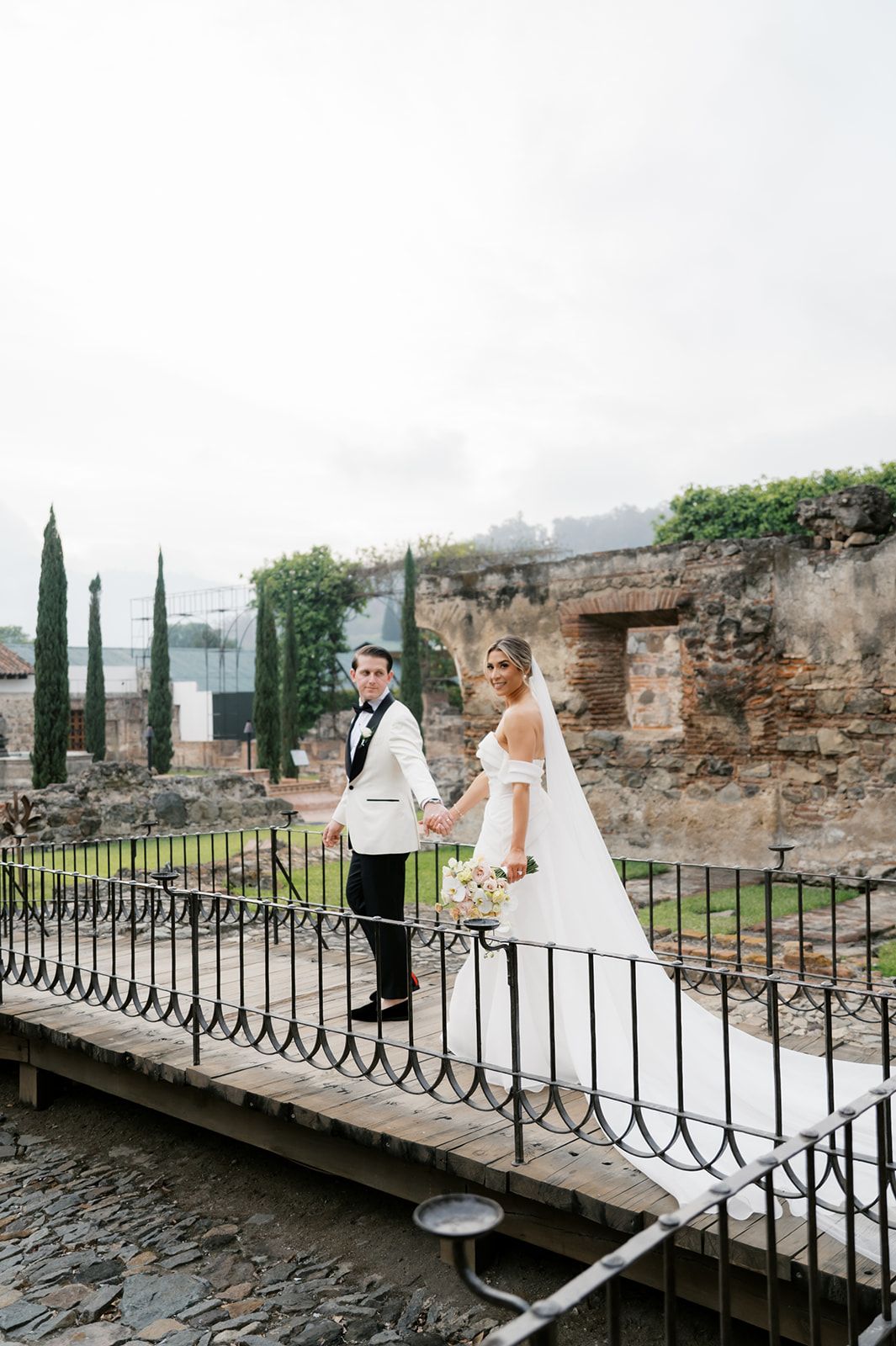 Bride and groom hold hands on a bridge at a historic stone venue, bride in off-shoulder gown with veil.