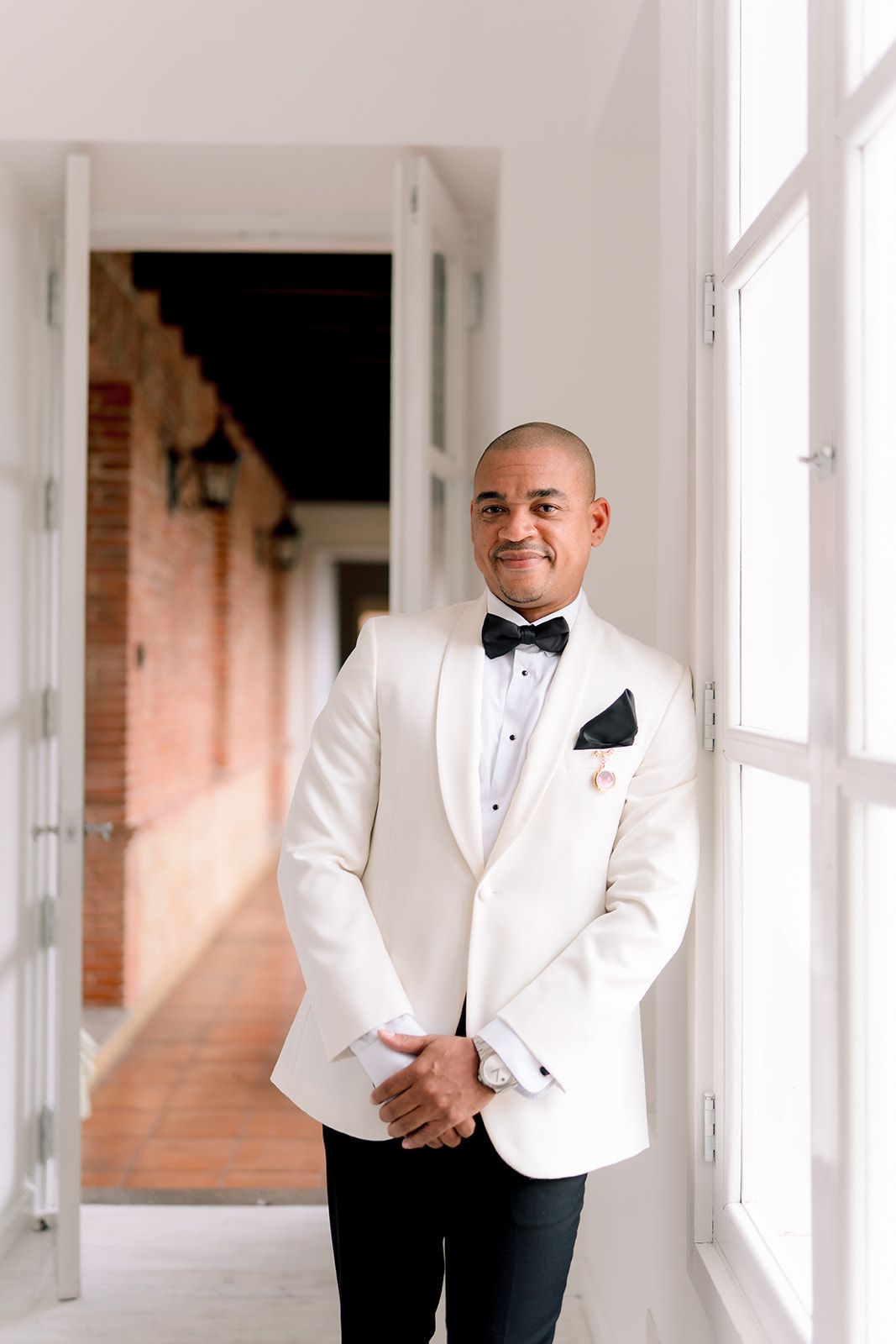 Smiling groom in white tuxedo jacket with black bow tie and pocket square stands by bright window at wedding venue.
