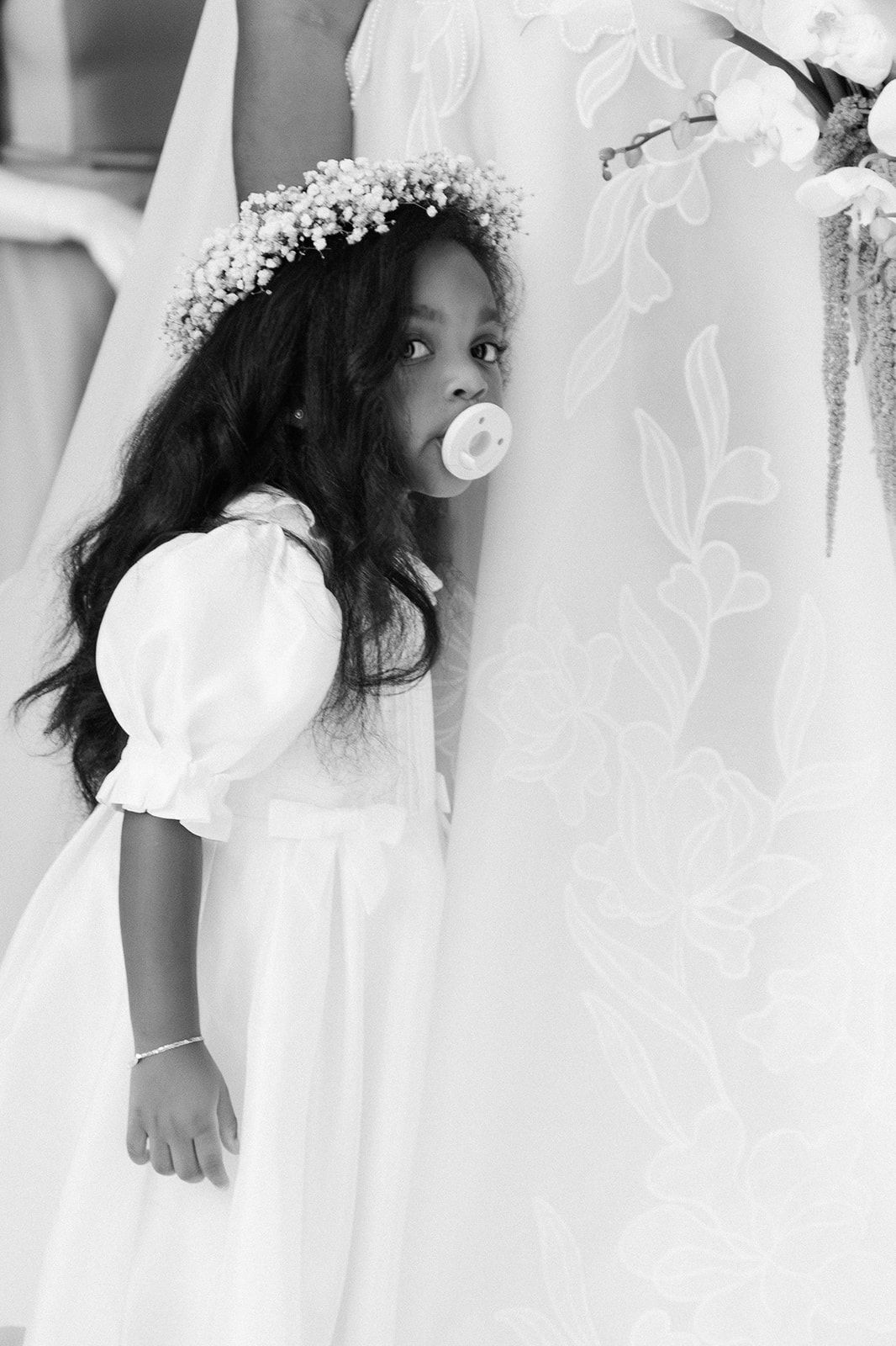 Black and white photo of a young flower girl in white dress with floral crown and pacifier, standing beside a bridal gown.