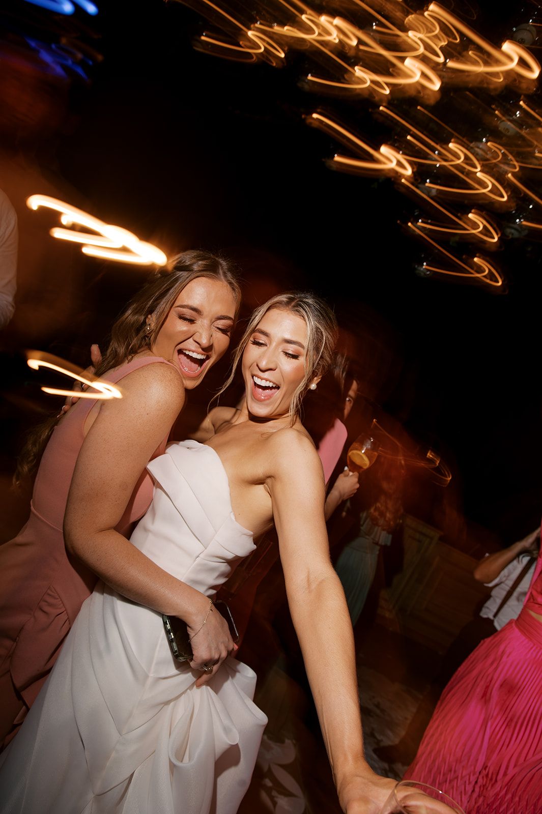 Two joyful women dancing at a wedding reception, one in white bridal gown, with bokeh lights in background.
