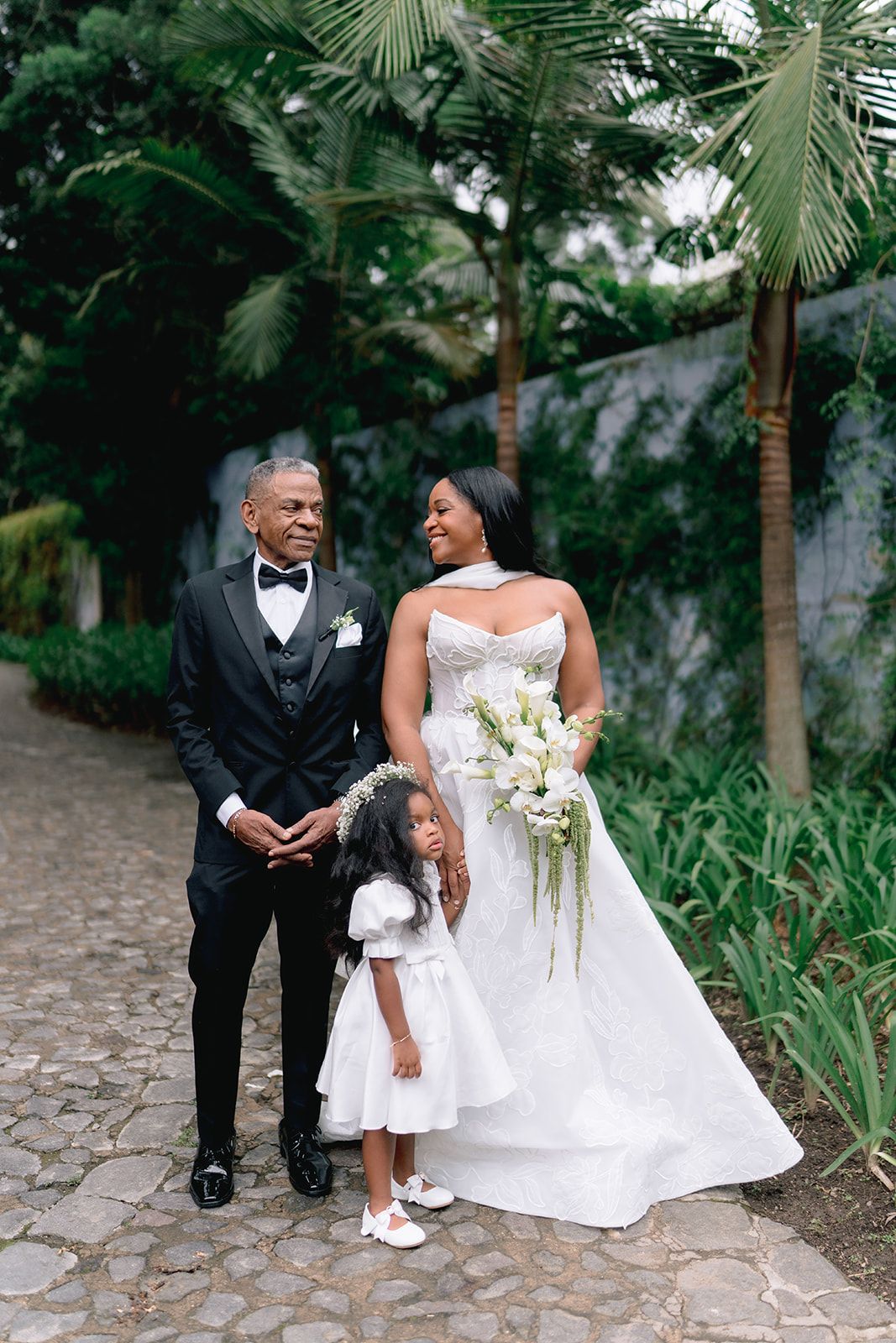 Bride and groom in elegant wedding attire pose with flower girl on stone path surrounded by lush tropical greenery.