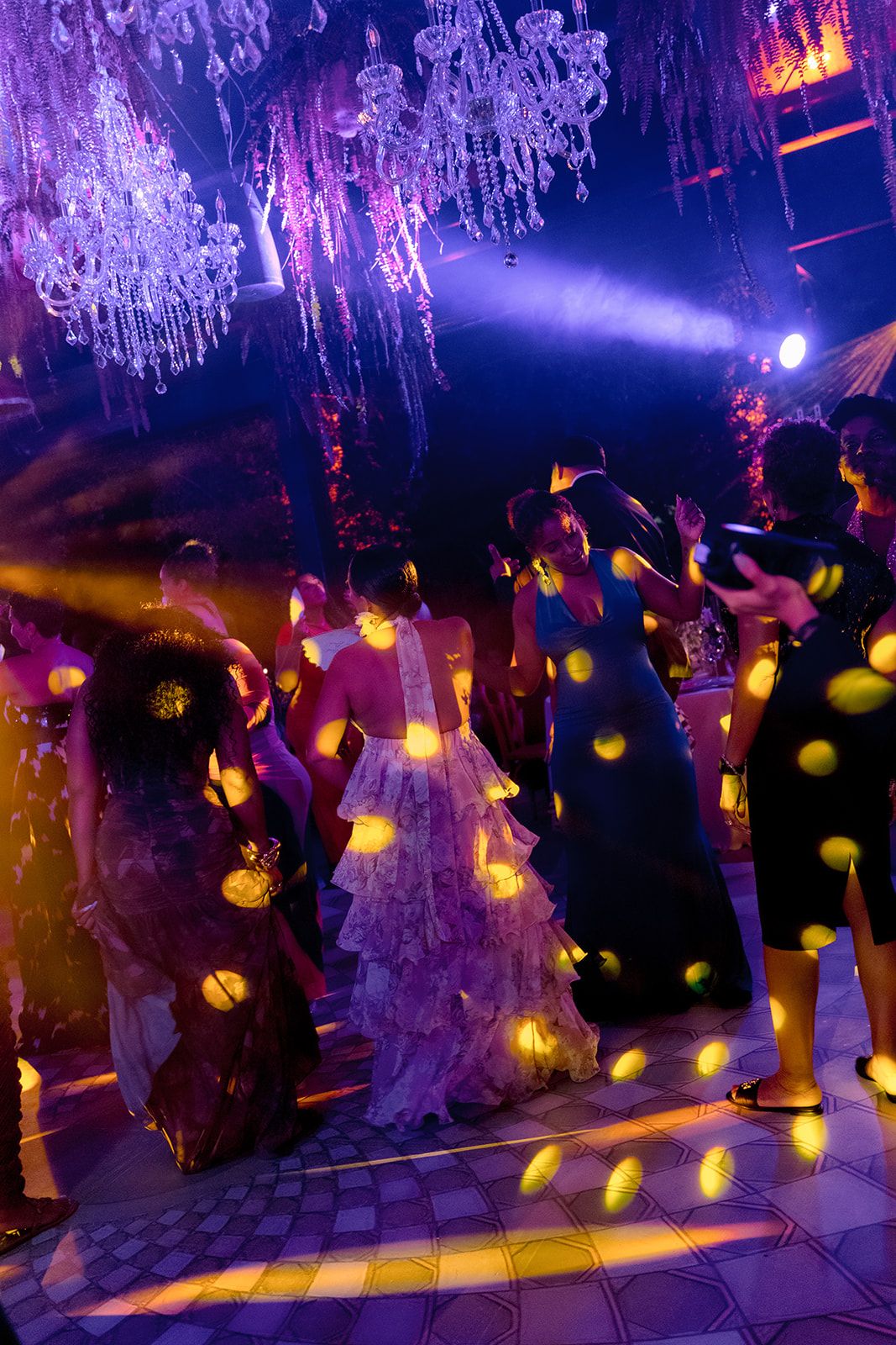 Elegant wedding reception dance floor with crystal chandeliers, purple lighting, and guests dancing amid yellow confetti.