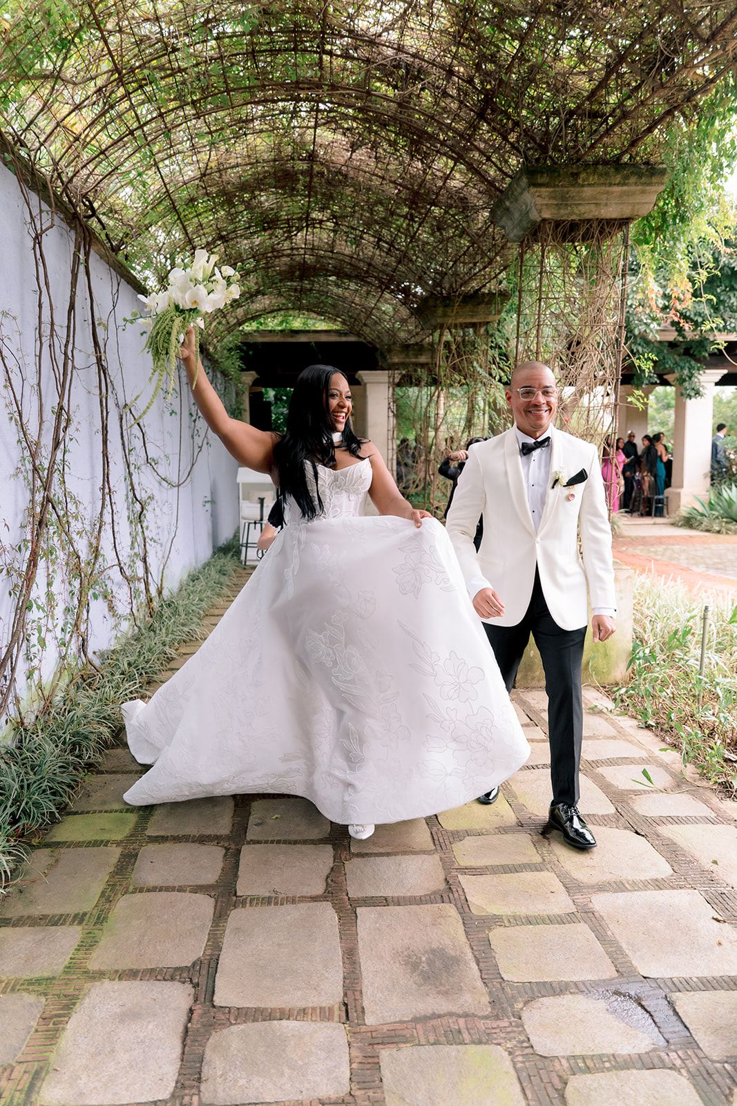 Joyful newlyweds walk under a vine-covered pergola, bride in a white ballgown holding bouquet aloft, groom in white tuxedo jacket.