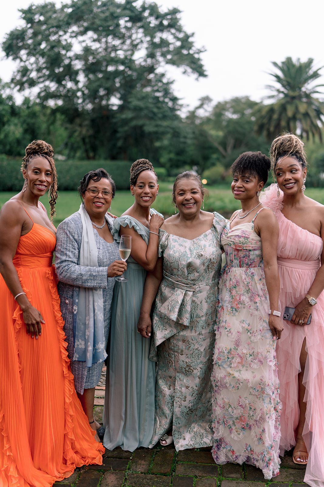 Six women in elegant formal gowns pose together outdoors at a garden wedding celebration.