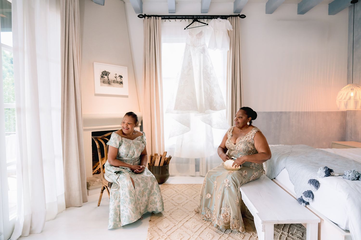 Two bridesmaids in matching floral gowns prepare in an elegant white bridal suite with a wedding dress hanging in the background.