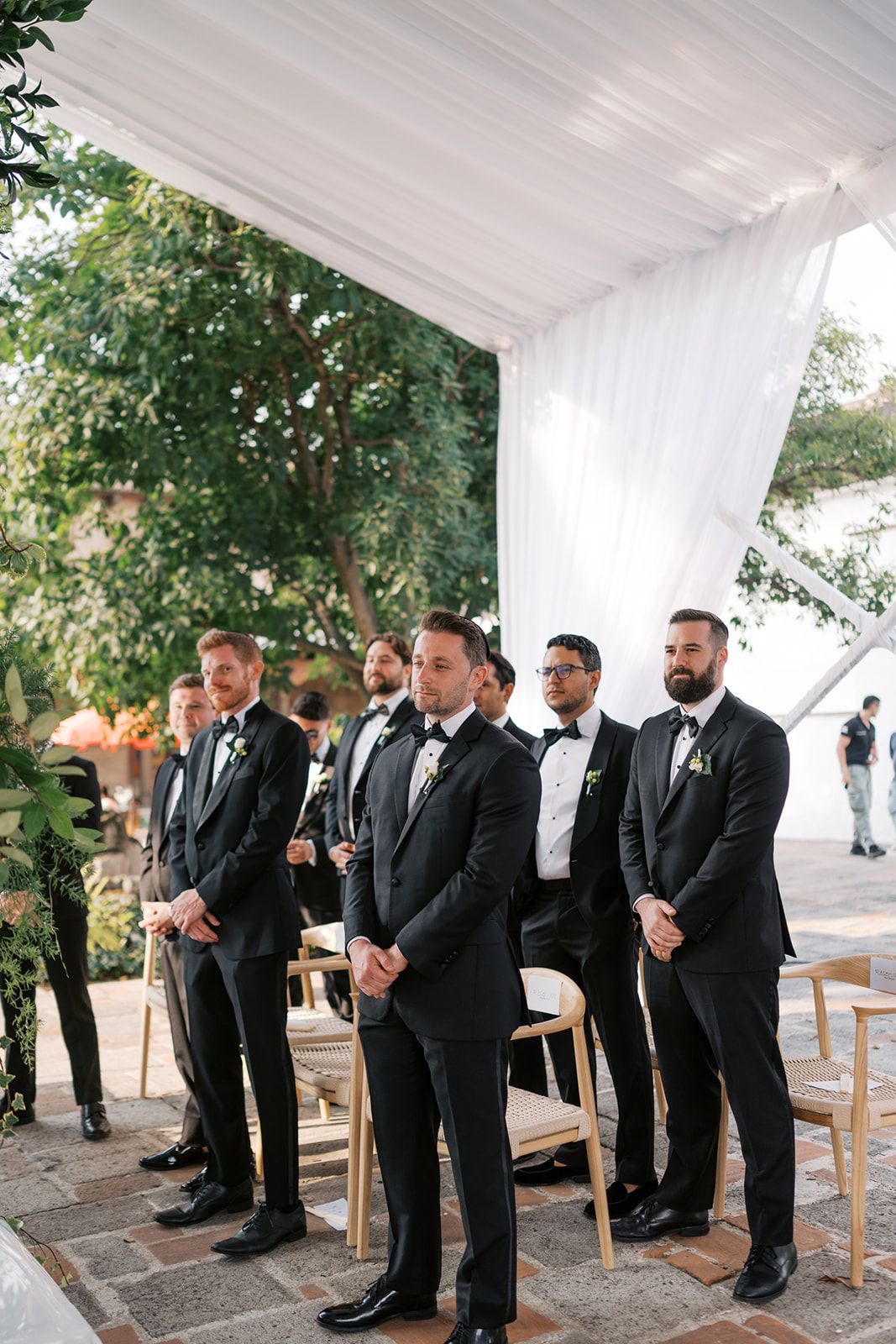 Groomsmen in black tuxedos with boutonnieres stand at an outdoor wedding ceremony under a white draped tent.
