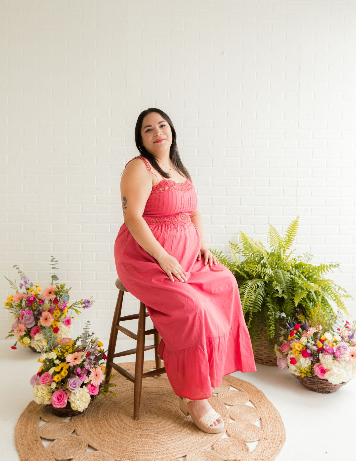 A woman in a coral pink maxi dress sits on a wooden stool surrounded by colorful flowers in a bright studio.