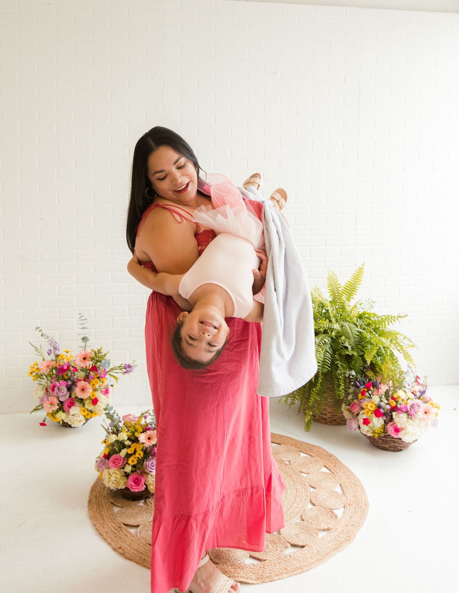 A joyful mother in a pink maxi dress playfully holds her laughing child upside down in a bright floral studio.