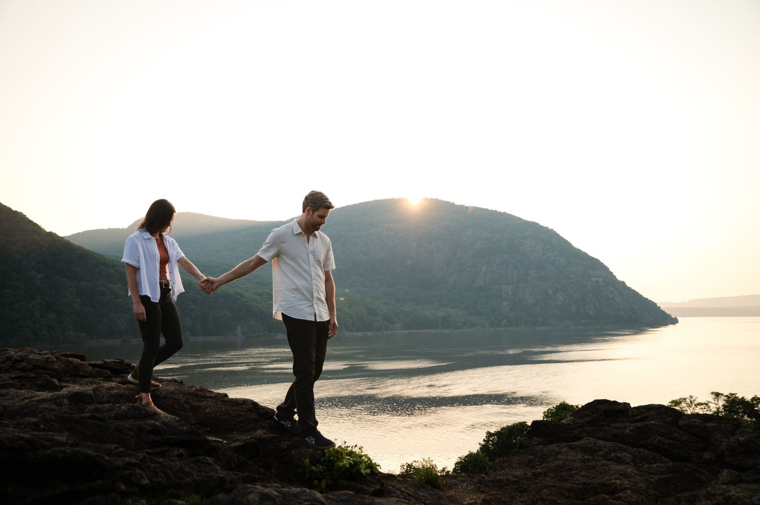 Two people in white shirts hold hands while walking along a rocky cliff overlooking a serene lake at sunset.