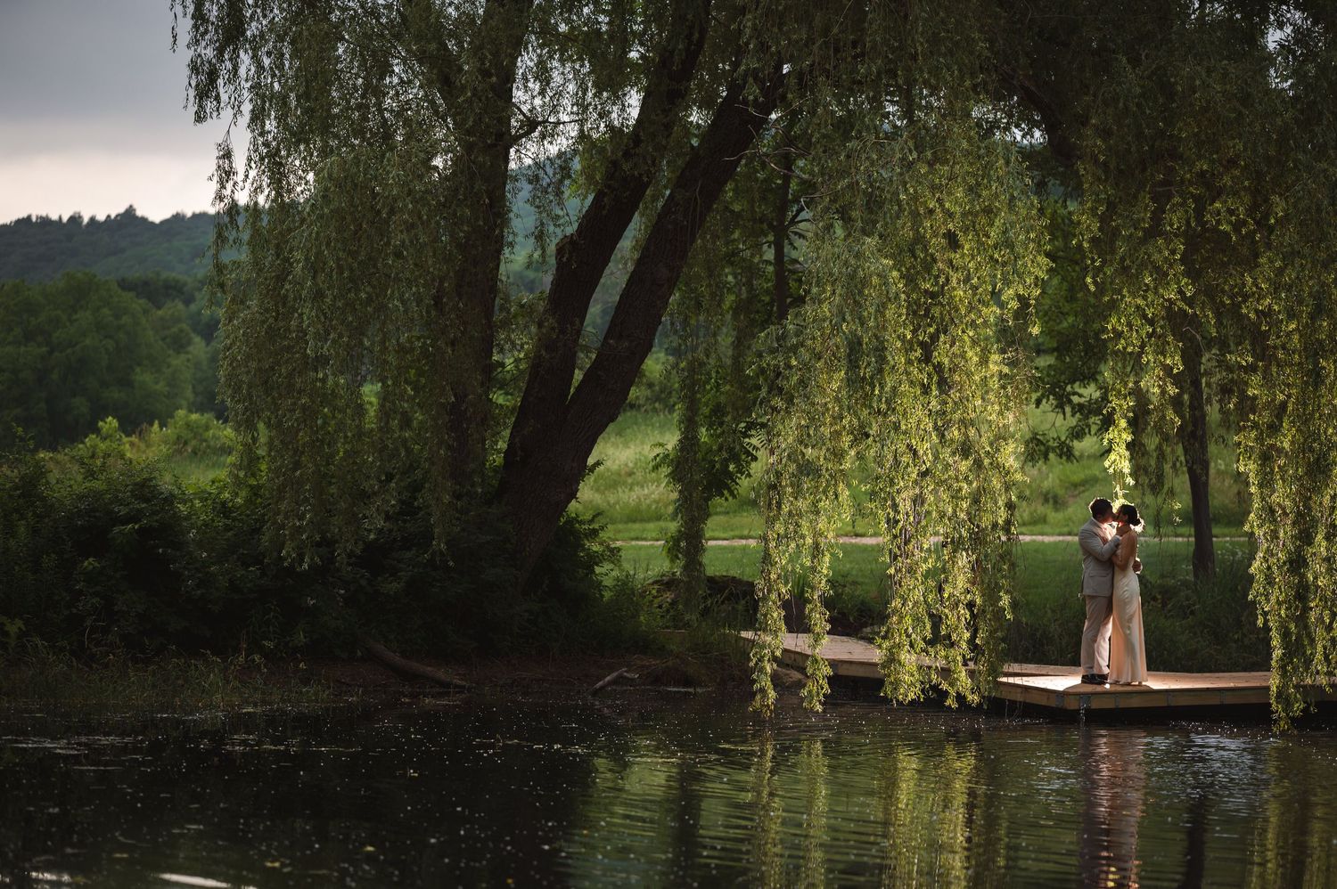Wedding couple kiss, silhouetted under weeping willow tree with reflection in water during golden hour at Ruins at Sassafras, Hudson Valley.