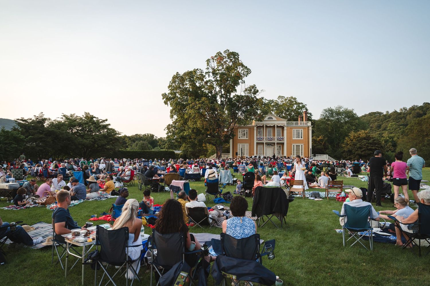 Large group of people sitting in lawn chairs during garden party at dusk on a grassy field at Boscobel in Garrison, NY in Hudson Valley