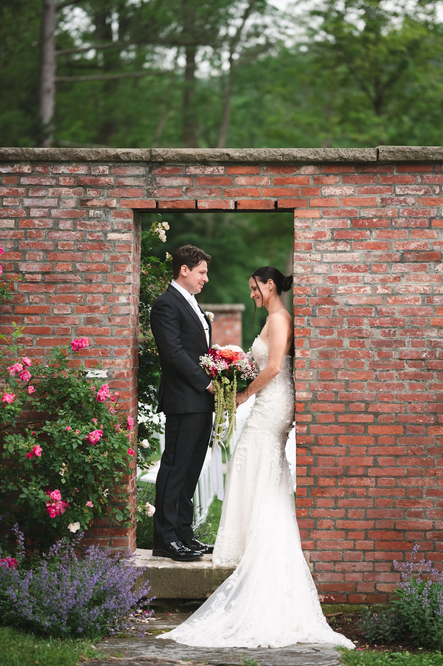 Newlyweds stand against a brick garden wall adorned with beautiful pink roses and purple flowers in romantic pose at The Ruins at Sassafras.