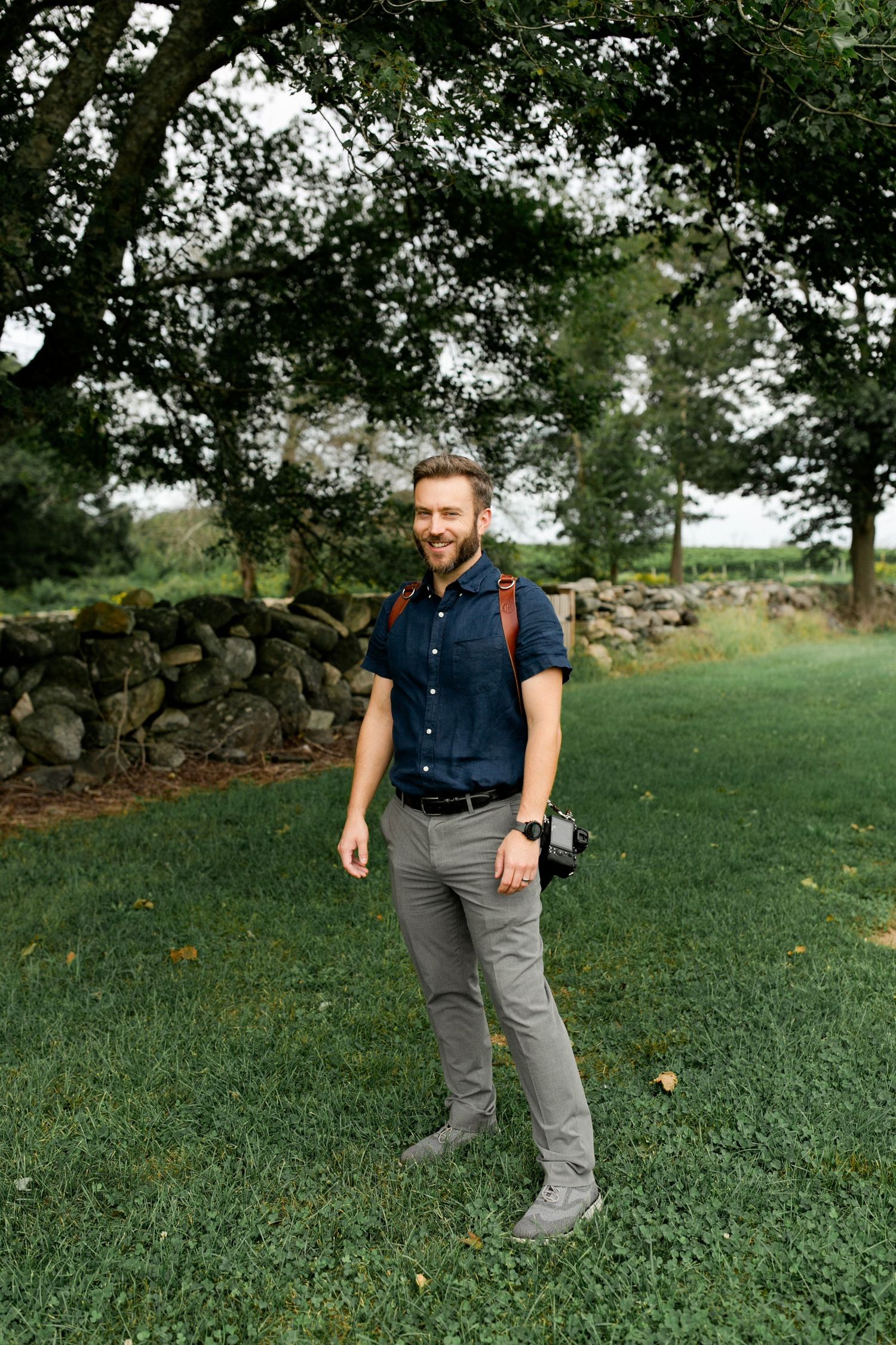 Matthew Rigby smiling with cameras on leather straps at an outdoor Hudson Valley wedding with stone wall, trees, and hills in background.