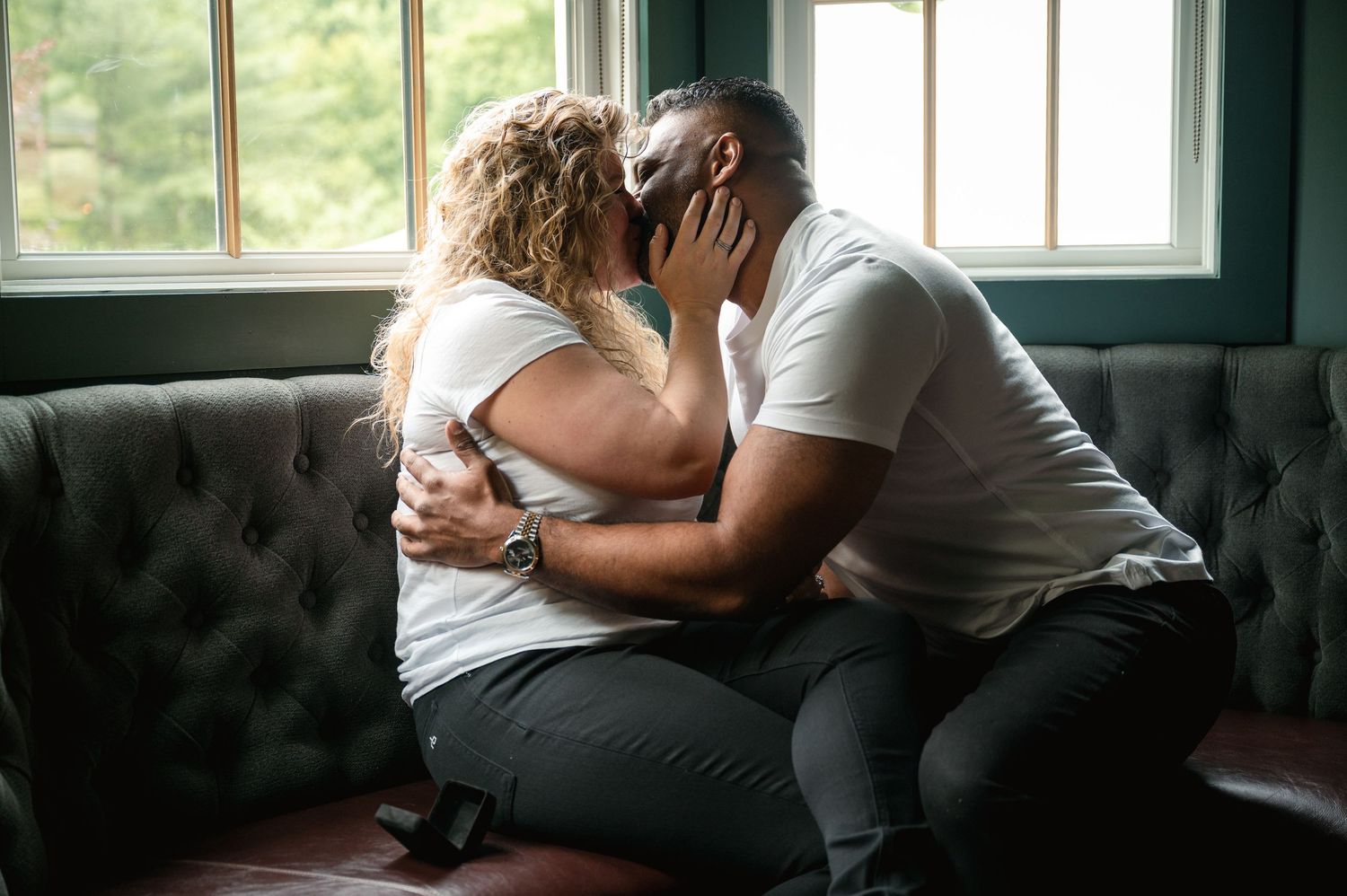 Couple lounging together on a tufted leather couch beneath bright window light.