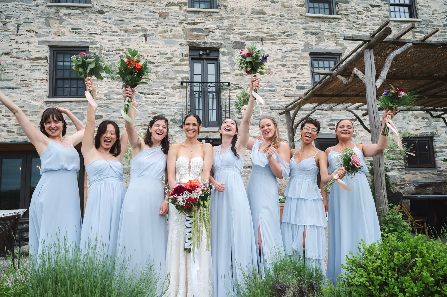 A row of bridesmaids and bride celebrate joyfully outside a historic rustic stone building at The Ruins at Sassafras in the Hudson Valley.