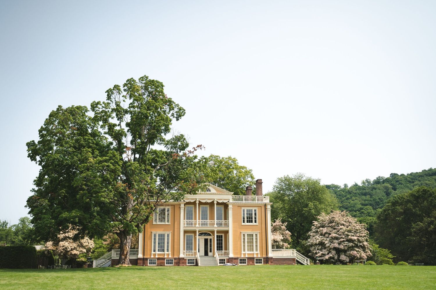 Elegant yellow antebellum mansion with white columns and wrap-around porch sits on a lush green lawn surrounded by trees.