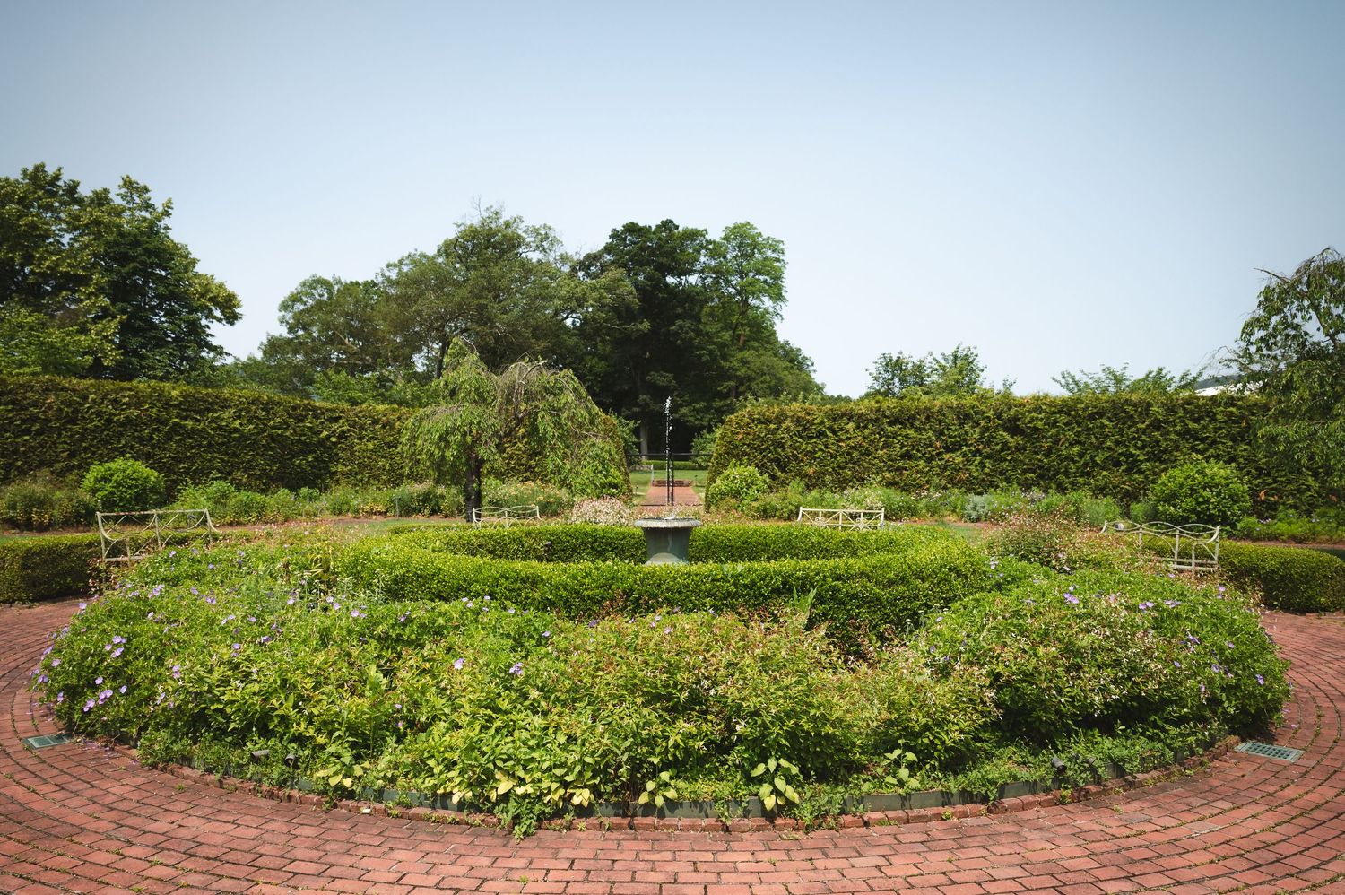 A circular brick garden path surrounding lush green ornamental garden with flowering plants and trees at Boscobel House in Garrison, NY.