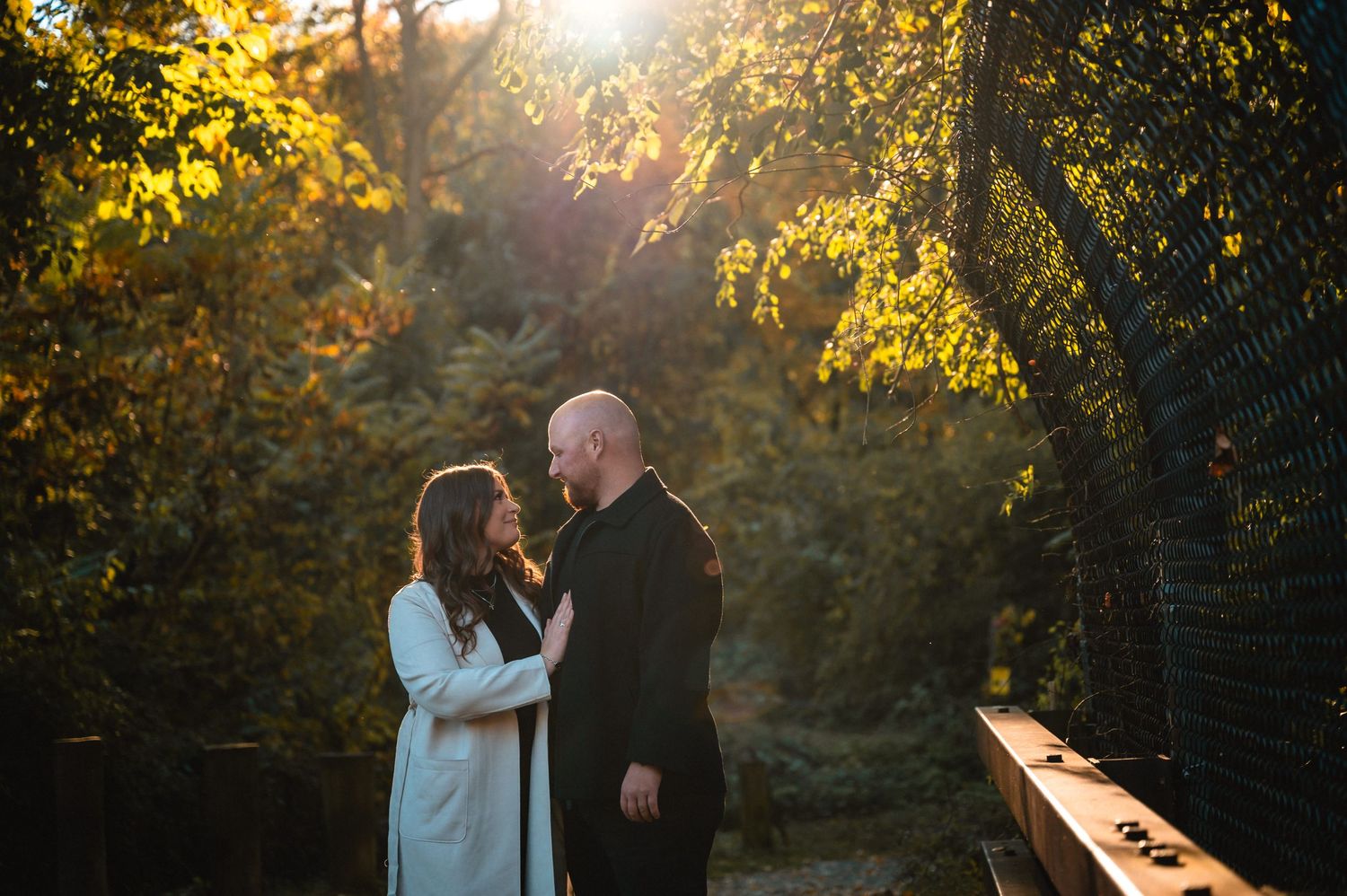 A couple shares an intimate moment on a bridge during golden hour sunset against an autumn forest backdrop.