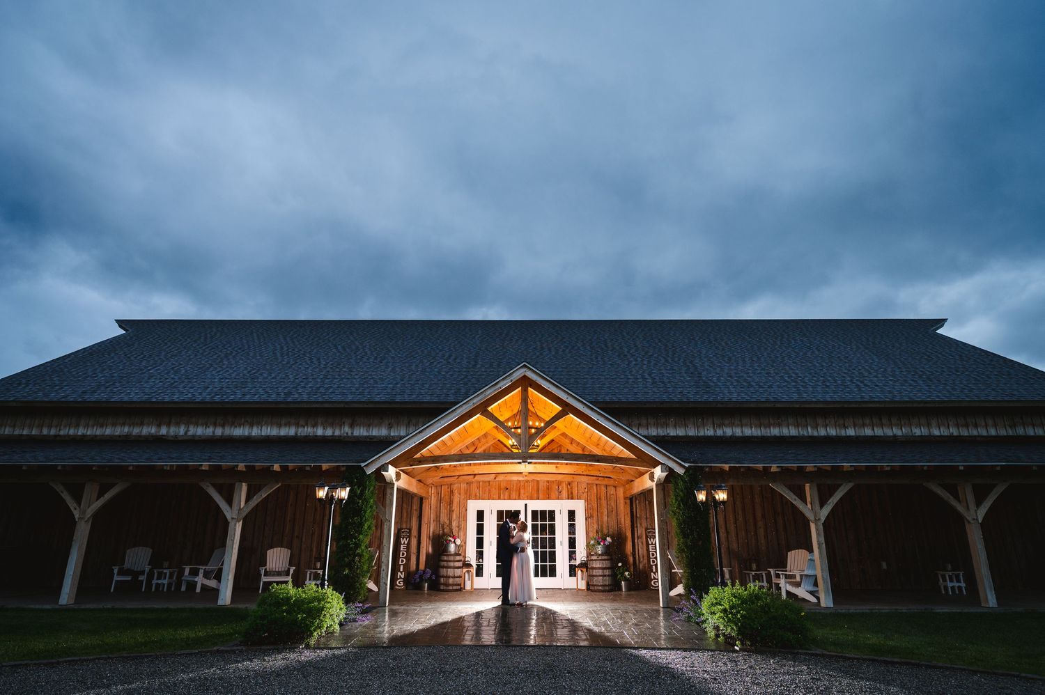 Illuminated rustic barn wedding venue entrance with glowing lights under dramatic stormy skies at dusk.
