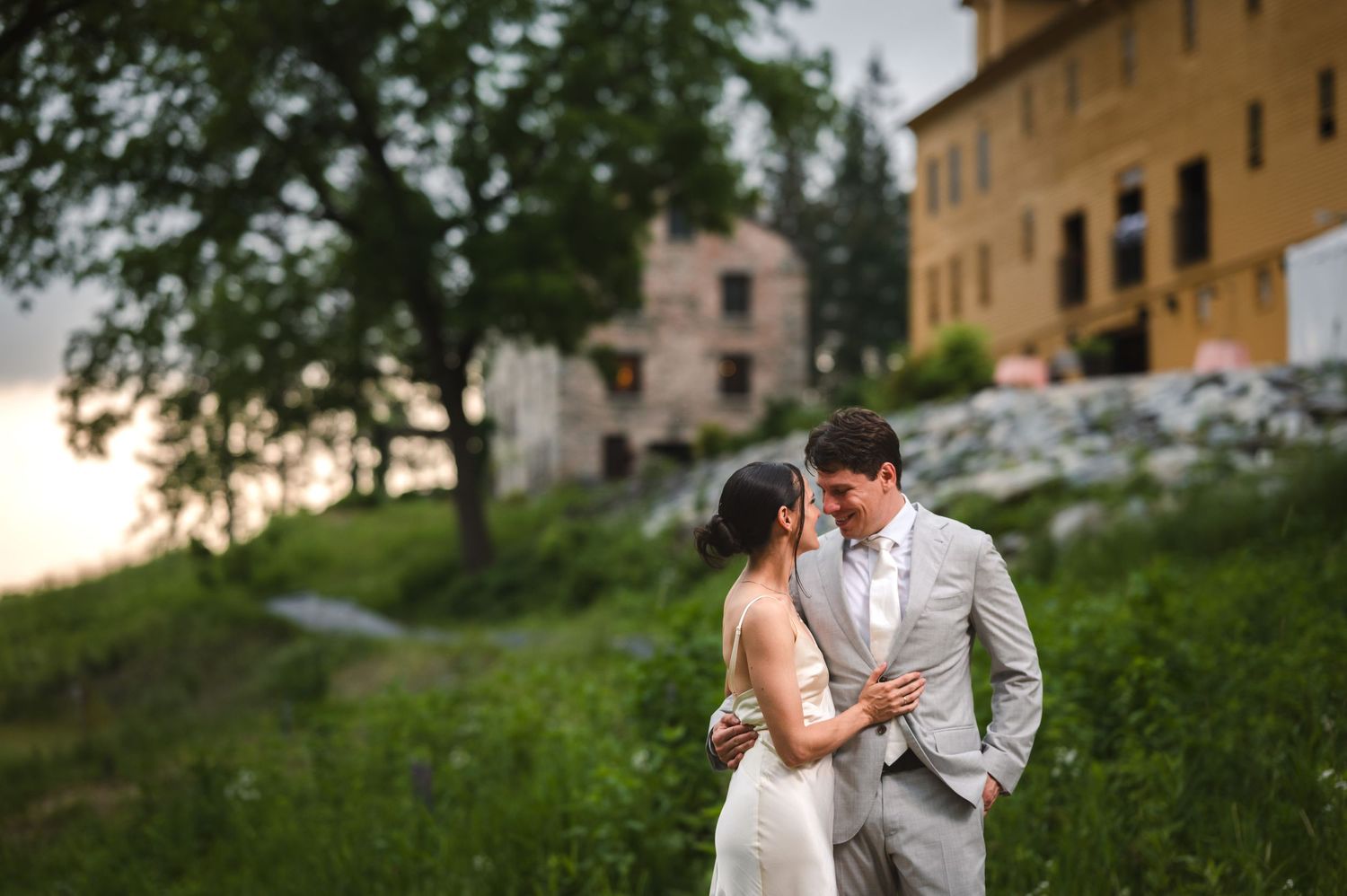 A romantic wedding couple embraces on a grassy hillside at sunset in front of rustic Italian style buildings at the Ruins at Sassafras NY.