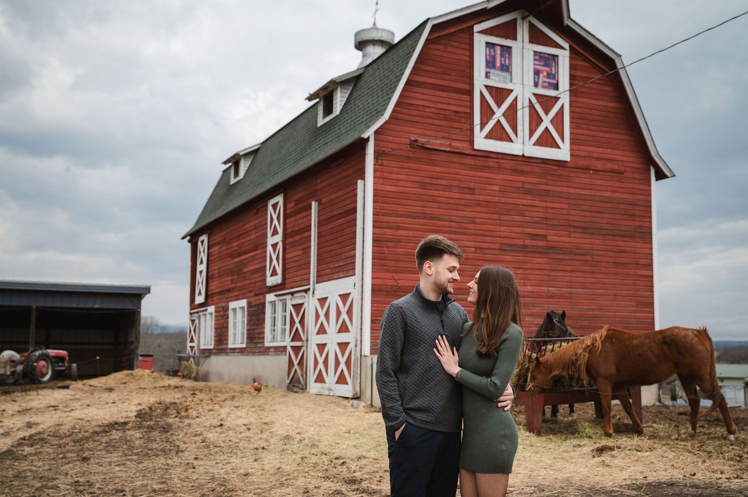 A couple embraces in front of a classic red barn with white trim and horses grazing in the background.