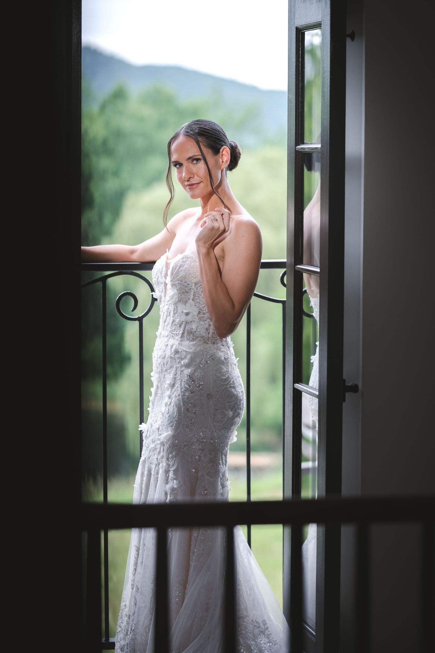 A bride in a white lace gown poses thoughtfully by a window with scenic views of green hills at The Ruins at Sassafras in the Hudson Valley.