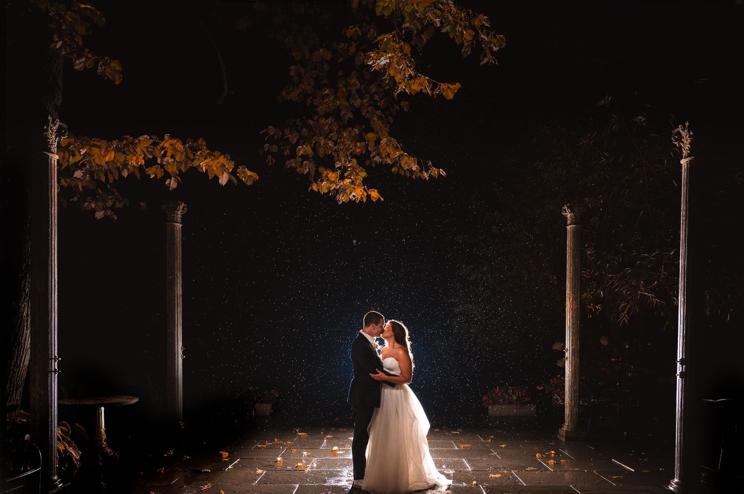 A cinematic portrait of couple kissing at night with atmospheric lighting and rain, on patio with pillars and fall trees in Hudson Valley.