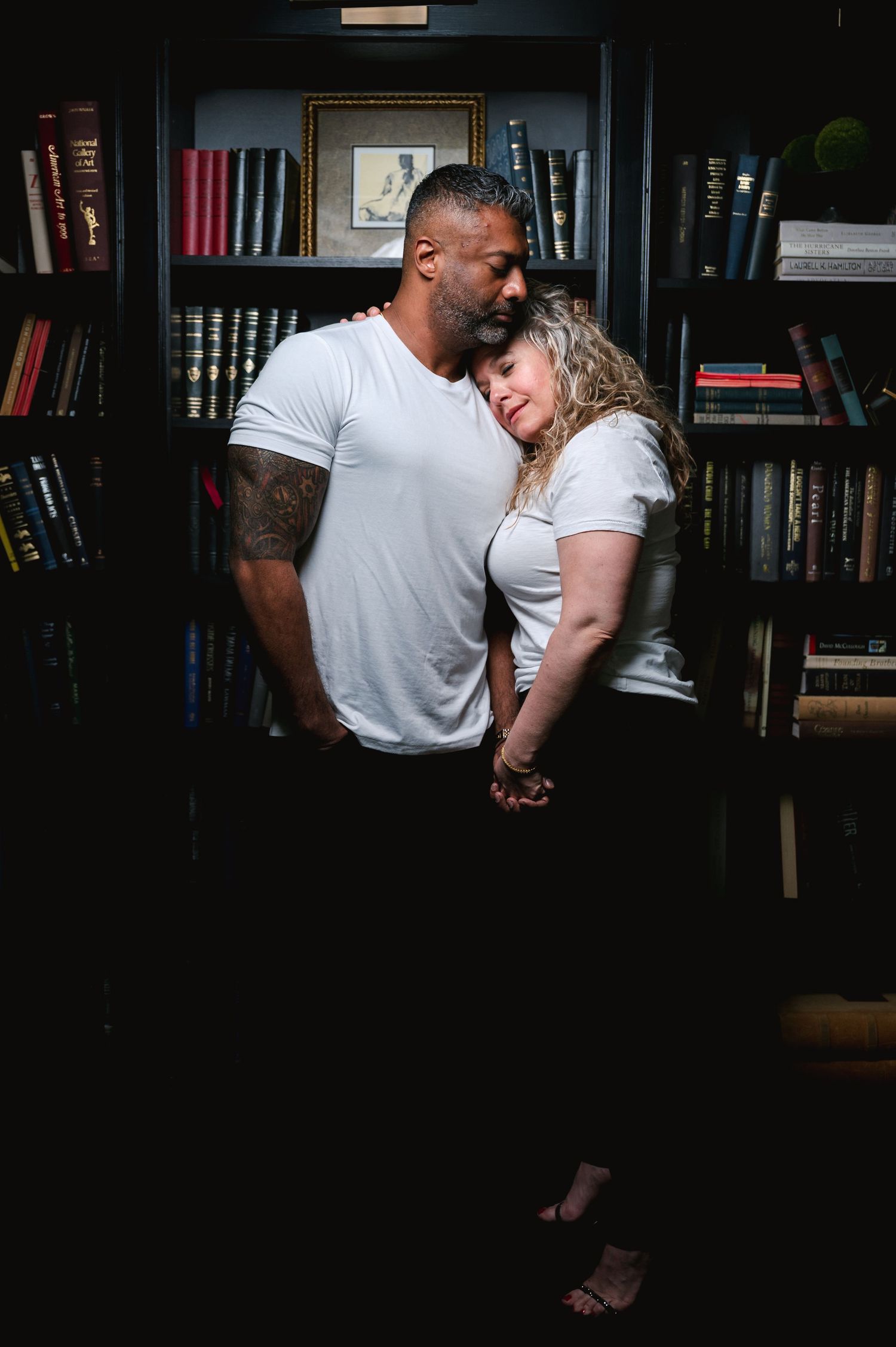 A couple embraces intimately while standing in front of a bookshelf in a dimly lit room.