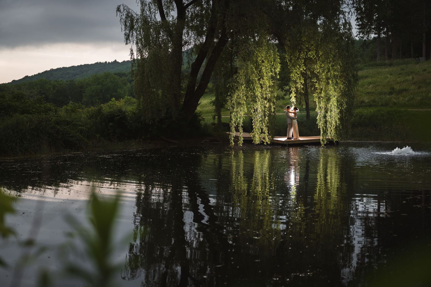 Silhouette of couple embracing under weeping willow tree with reflection in water during golden hour at Ruins at Sassafras, Hudson Valley.