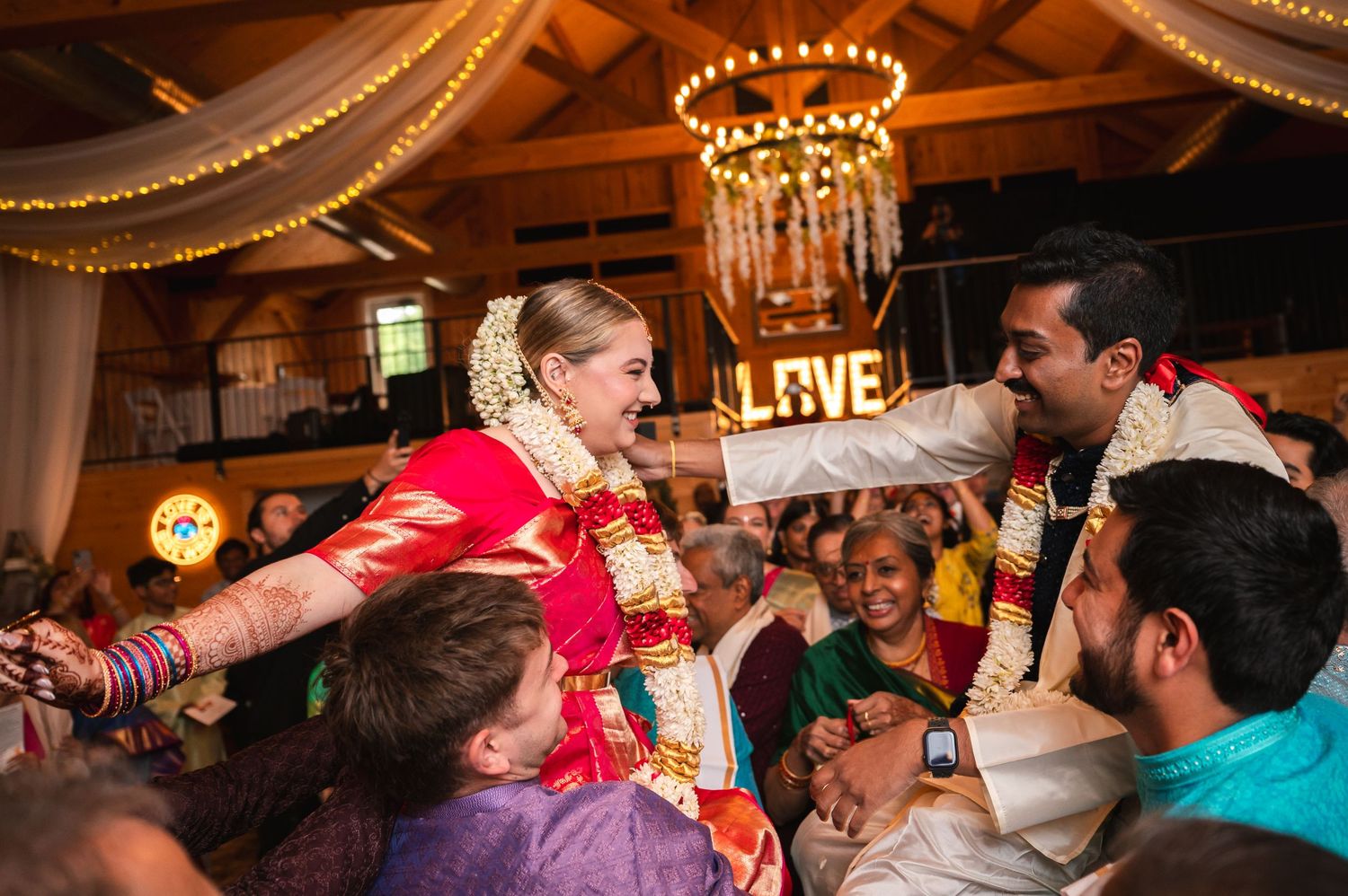 Bride and groom in Indian attire on guests shoulders in rustic barn with beams and chandelier at Fortune Valley Manor in Saugerties, NY.