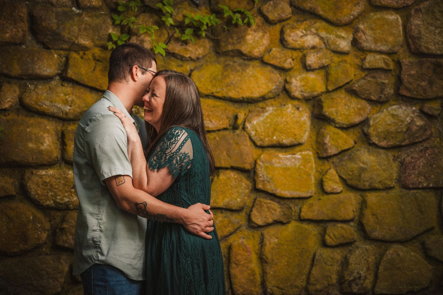 Two people share an intimate moment against a rustic stone wall background as sunlight creates a warm glow.