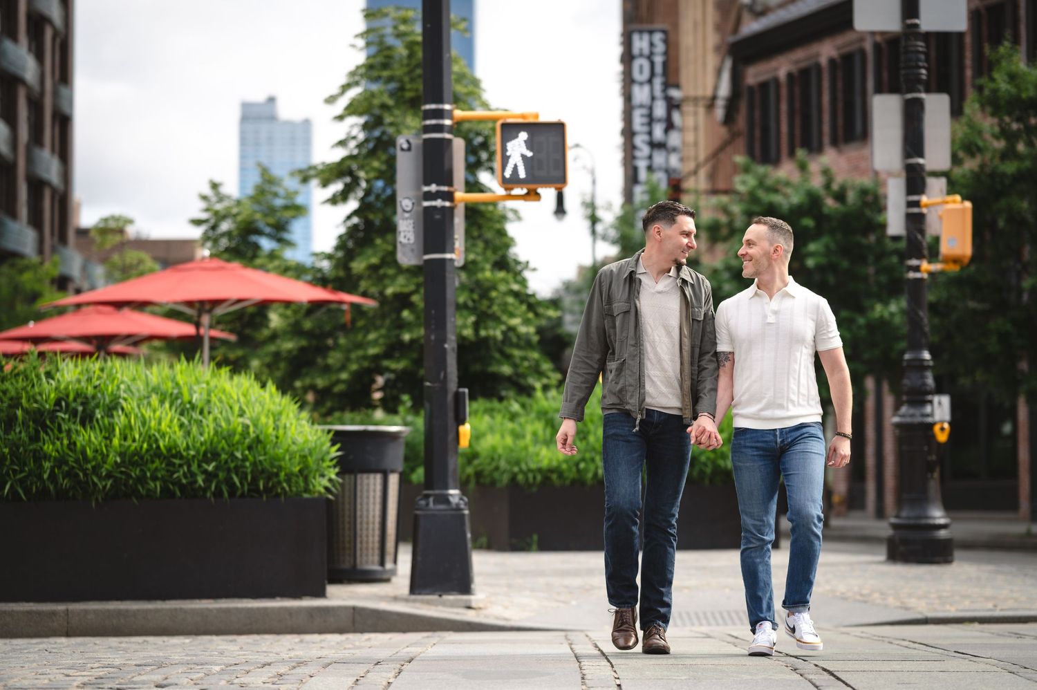 Two people walking together and smiling on an urban sidewalk with red umbrellas and city buildings in the background.