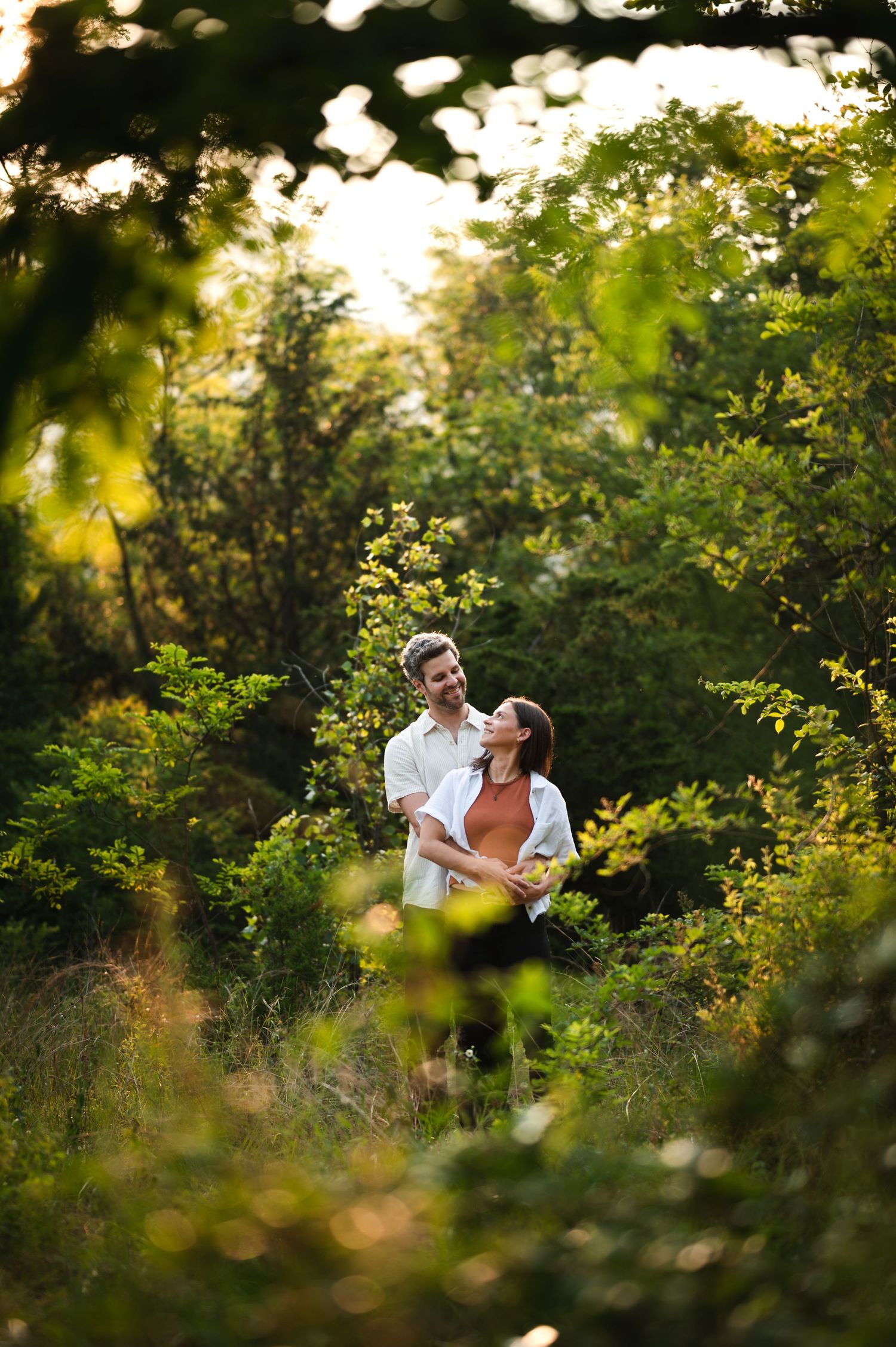 Two people share a romantic moment surrounded by lush greenery and golden sunlight in an outdoor setting.