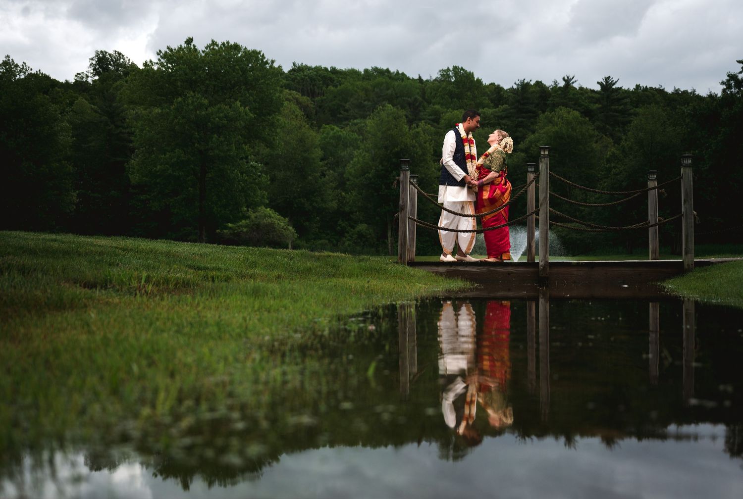 Couple in Indian wedding attire stands on a wooden bridge over a pond, reflected in the still water at Fortune Valley Manor in Hudson Valley