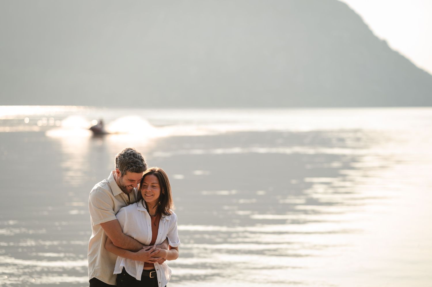 A couple walking on the beach at sunset shares an intimate moment while holding their baby under golden light.