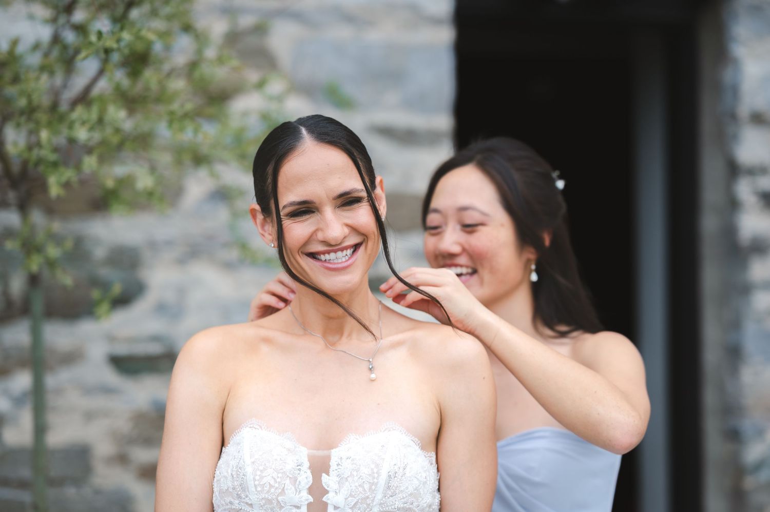 Bridesmaid helps bride put on necklace in front of stone wall, sharing candid laughs at The Ruins at Sassafras in the Hudson Valley.
