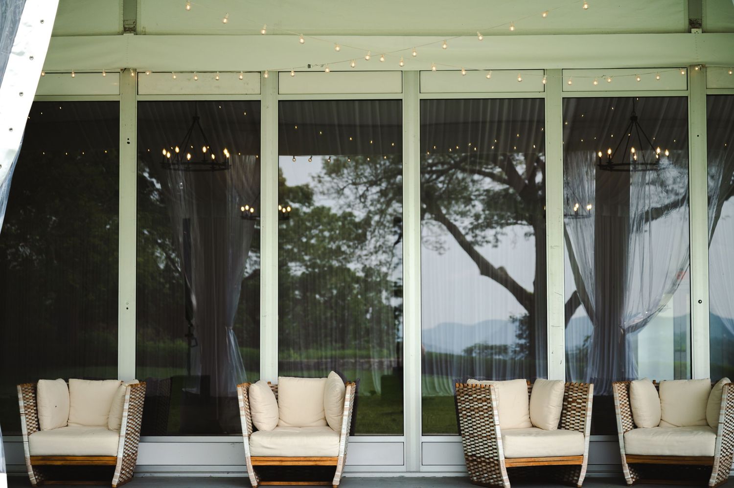 Modern wedding tent porch with cushioned seating against floor-to-ceiling windows overlooking water and trees at Boscobel in Garrison, NY.