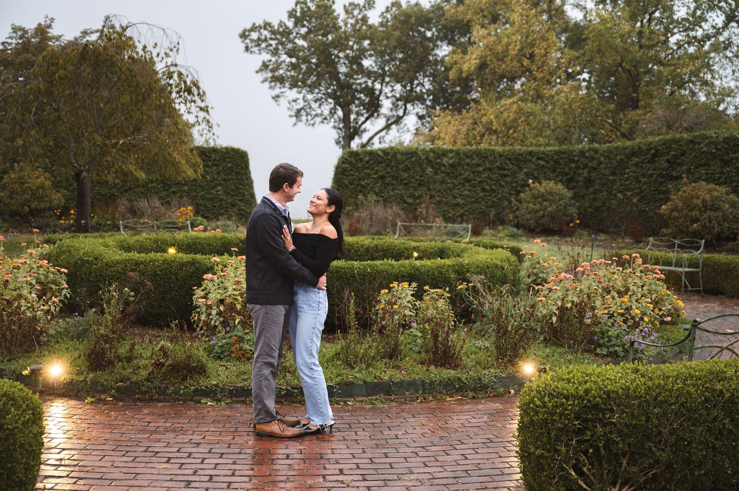 Newly engaged couple at a brick garden path surrounding lush green ornamental garden with flowering plants and trees at Boscobel House