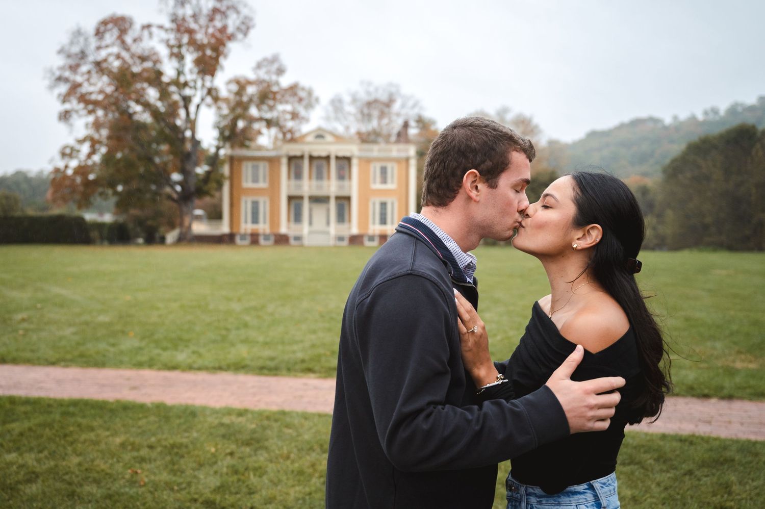 Couple kissing in front of mansion estate with white columns and lush green landscaping at Boscobel House in Garrison, NY in Hudson Valley.