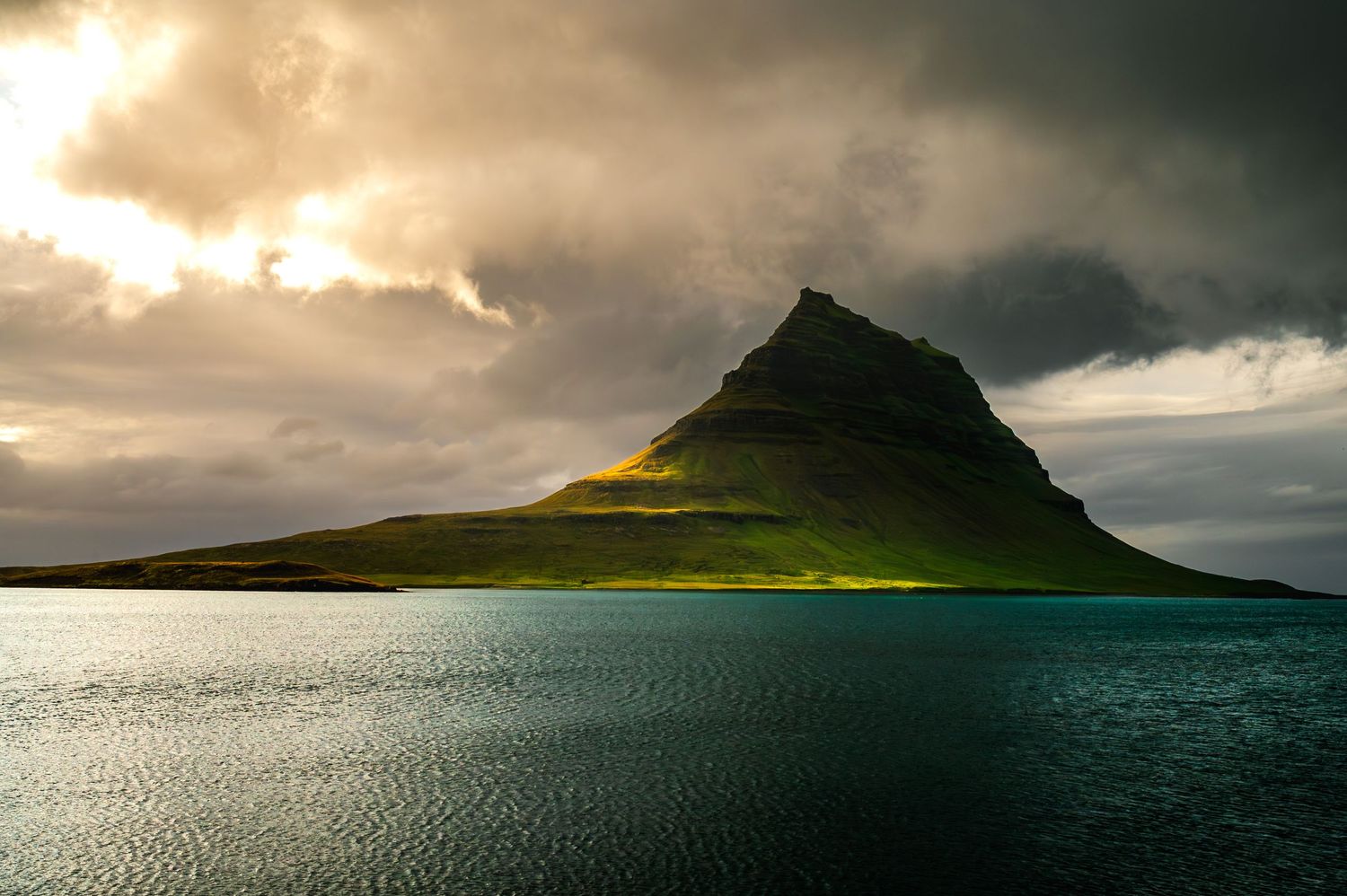 Dramatic sunlight breaks through stormy clouds over Kirkjufell mountain in Iceland, casting golden rays on water and green slopes.