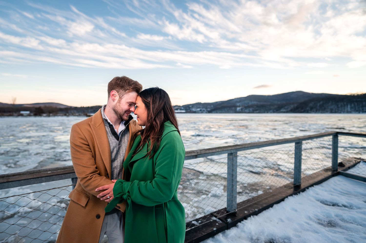 A couple in winter coats embraces on a snowy deck overlooking a frozen lake and mountains on a sunny day.