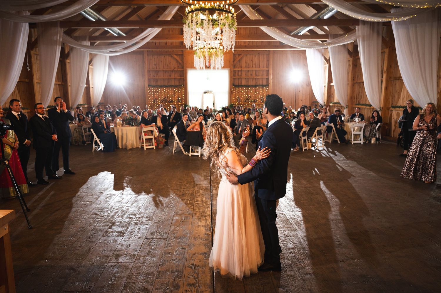 A couple shares their first dance in a rustic barn reception at Fortune Valley Manor, illuminated by warm lighting and surrounded by guests.