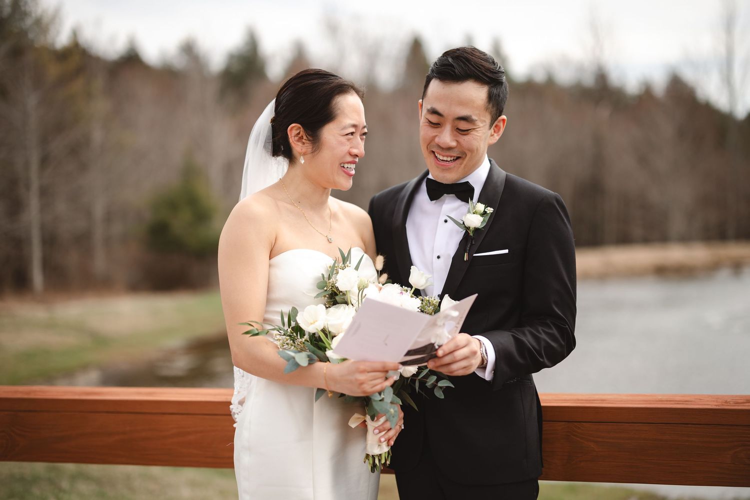 A couple in formal wedding attire shares a joyous candid moment reading vows to each other on bridge at Windham Manor, Hudson Valley, NY.