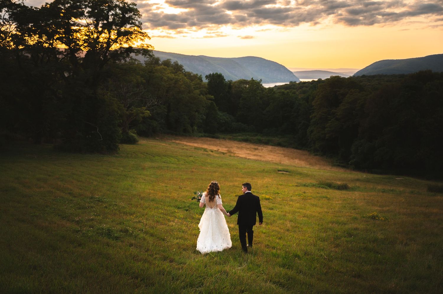 Wedding couple walks hand in hand in a meadow at sunset towards silhouetted mountains and trees at The Garrison, Garrison NY. Hudson Valley.