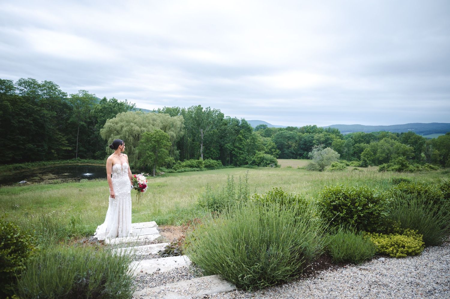 Bride stands on a stone path overlooking rolling hills and gardens on a cloudy summer day at The Ruins at Sassafras in the Hudson Valley.