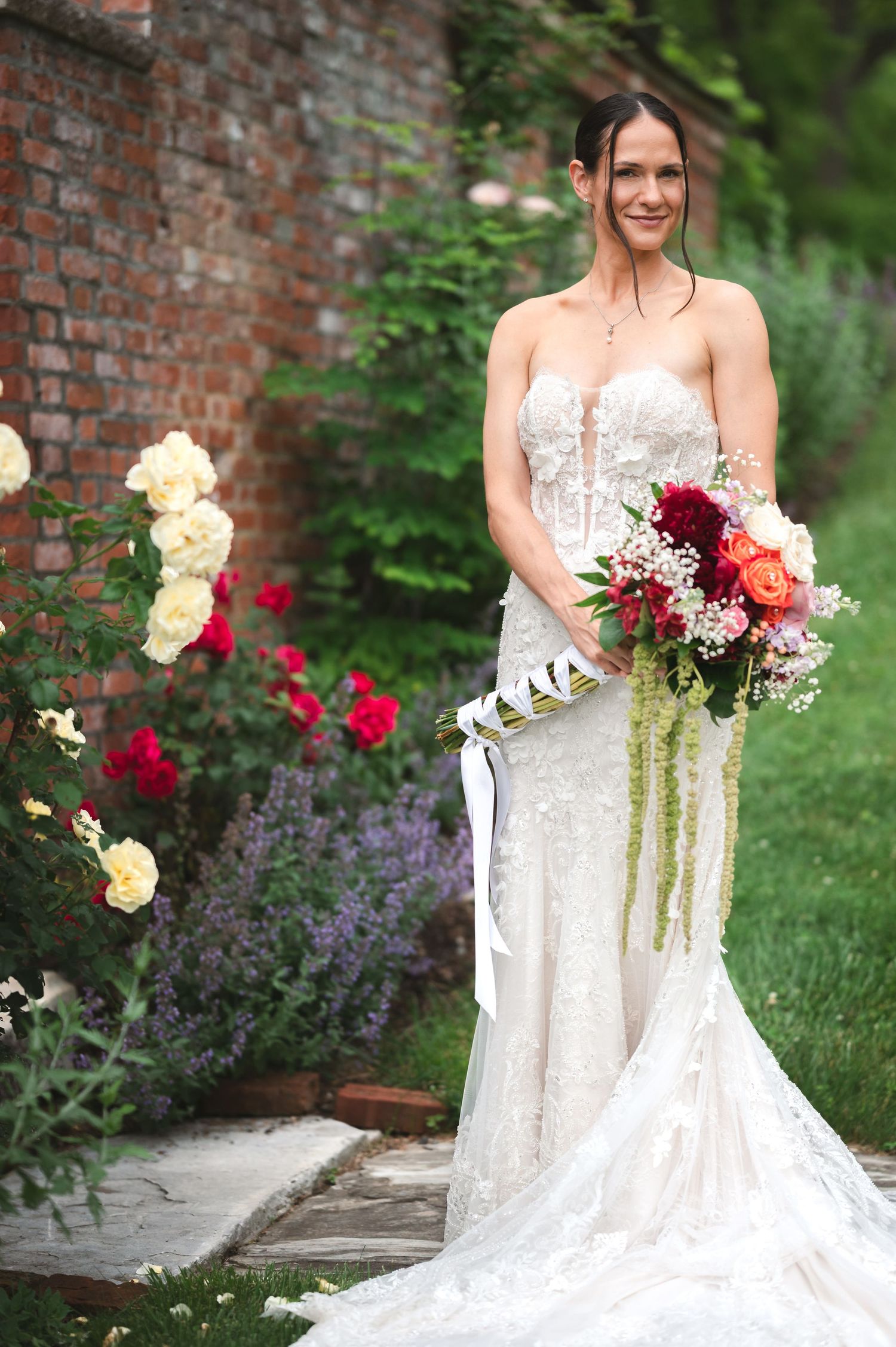 A bride in a white lace wedding gown holds a burgundy and cream bouquet against a rustic brick garden backdrop at The Ruins at Sassafras NY.