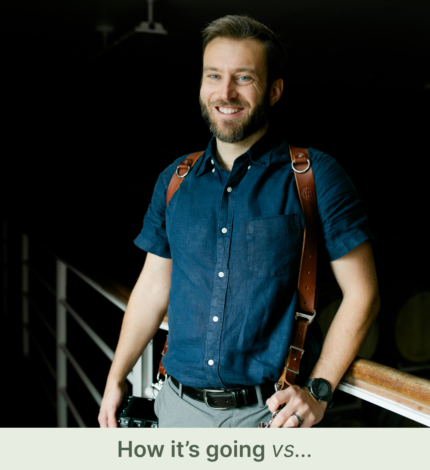 Matthew Rigby smiling with cameras on leather straps in Hudson Valley winery wedding with wooden railing and barrel room in the background.